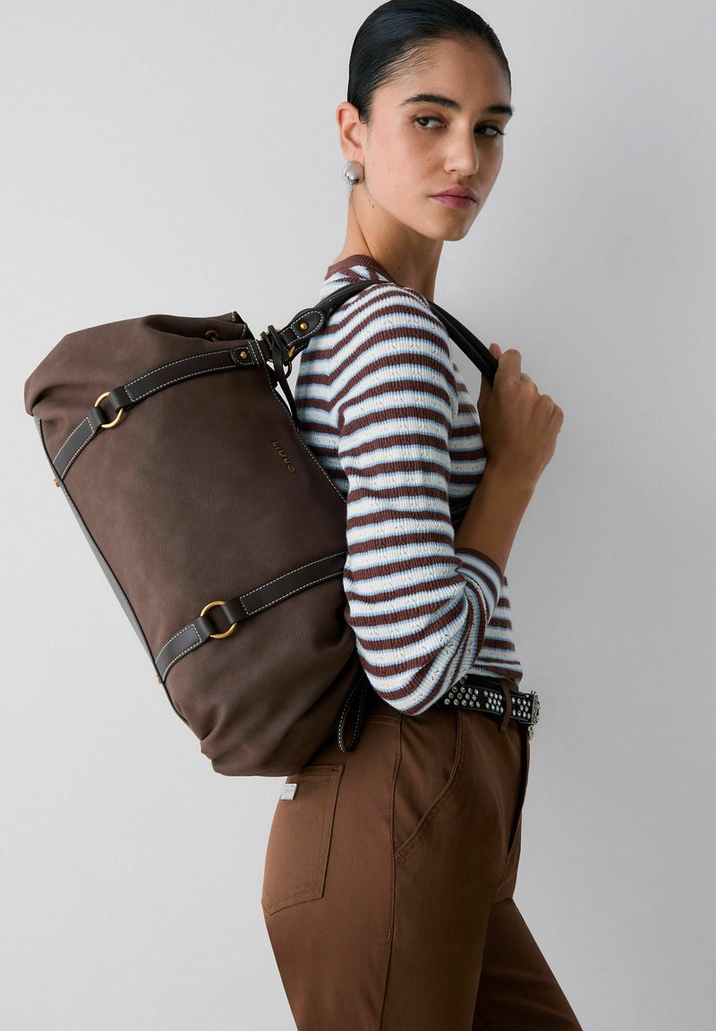 Femme aux cheveux tirés en arrière portant un pull rayé et un pantalon marron, avec un grand sac à dos en cuir marron foncé porté sur une épaule.