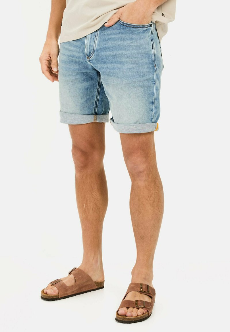 Shorts en denim bleu clair avec des revers, présentant un design classique à cinq poches et un léger dégradé. Sandales en daim marron avec boucles.