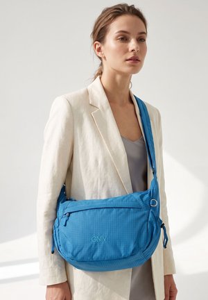 Jonge vrouw met een crèmekleurige blazer die een grote blauwe crossbodytas draagt met een rits aan de voorkant en GMV-logo.