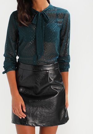 Femme portant un chemisier transparent vert foncé à pois avec un nœud au col et une mini-jupe en cuir noir brillant, bras relâchés le long du corps.