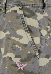 Kamufliažinės kelnių modelis žaliais ir smėlio atspalviais, su rožiniu drugelio logotipu šalia kišenės ir standartine džinsinio audinio siuvimo technologija.