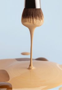 Pinceau de maquillage avec un fond de teint liquide beige dégoulinant sur une surface lisse sur fond bleu clair.