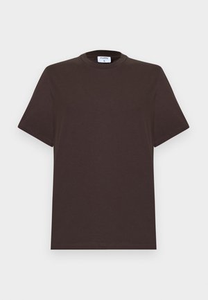 Femme aux cheveux foncés portant un t-shirt noir, un pantalon beige clair à jambes larges et des sandales marron, debout les mains dans les poches sur fond blanc.