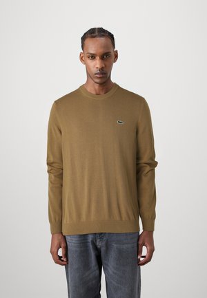 Brown knit sweater with long sleeves, round neckline, ribbed cuffs, and hem. Features a small green logo on the chest. Worn with gray jeans.