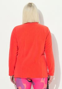 Punainen fleece-takki, jossa on pitkät hihat, tiukka malli, pehmeä rakenne ja hienovaraiset saumat, yhdistettynä monivärisiin kuviollisiin legginsseihin.