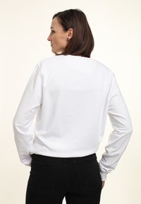 Felpa bianca a maniche lunghe con colletto a girocollo e polsini a costine, abbinata a jeans neri attillati, vista da dietro. Tessuto dalla superficie liscia.