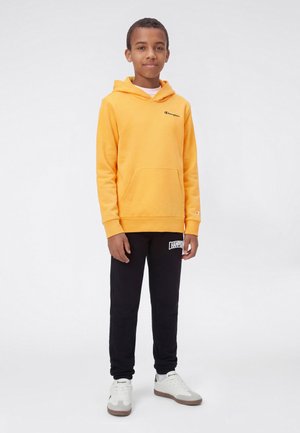 Garçon debout portant un sweat à capuche jaune Champion, un pantalon de survêtement noir Champion et des baskets blanches sur un fond blanc.
