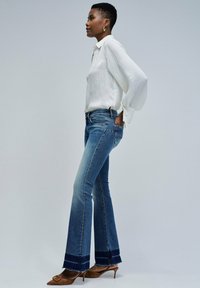 Jeans de mezclilla acampanados en azul medio con un dobladillo deshilachado y un puño oscuro en contraste, combinados con una blusa blanca de manga larga con textura y tacones marrones.