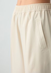 Pantaloni color crema realizzati in un tessuto leggero con un sottile motivo a griglia, dotati di una cintura elasticizzata e una tasca laterale.