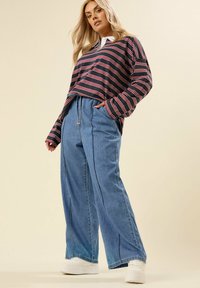 Donna bionda che indossa una polo a maniche lunghe a righe, jeans blu a gamba larga con coulisse e sneakers bianche con piattaforma, in piedi con una mano in tasca.