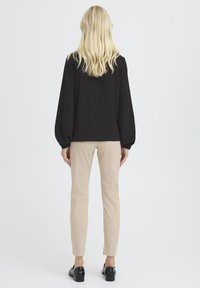 Blond kvinde, der står med ryggen til iført en sort langærmet bluse, beige slanke bukser og sorte loafers foran en ensfarvet baggrund.