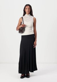 Krämfärgad, ribbad ärmlös topp med spetsdetaljer, kombinerad med en fladdrande svart maxikjol. Modellen håller en liten brun handväska och bär svarta ballerinaskor.