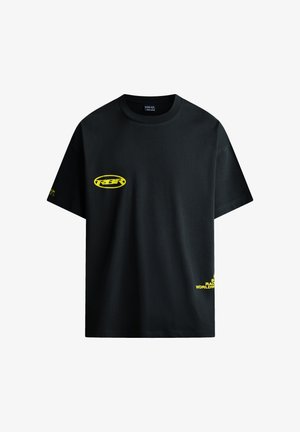 T-shirt nera a maniche corte con piccolo logo ovale giallo "REBEL" sul petto, testo giallo vicino all'orlo inferiore e testo giallo sulla manica sinistra.
