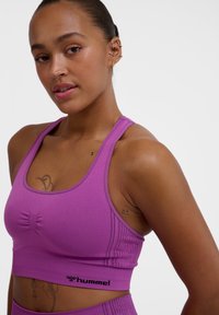 Fioletowy sportowy top w wersji crop z ribbowanej tkaniny, z wyciągniętym przodem i logo na dolnej krawędzi, zaprojektowany z myślą o komforcie i wsparciu.