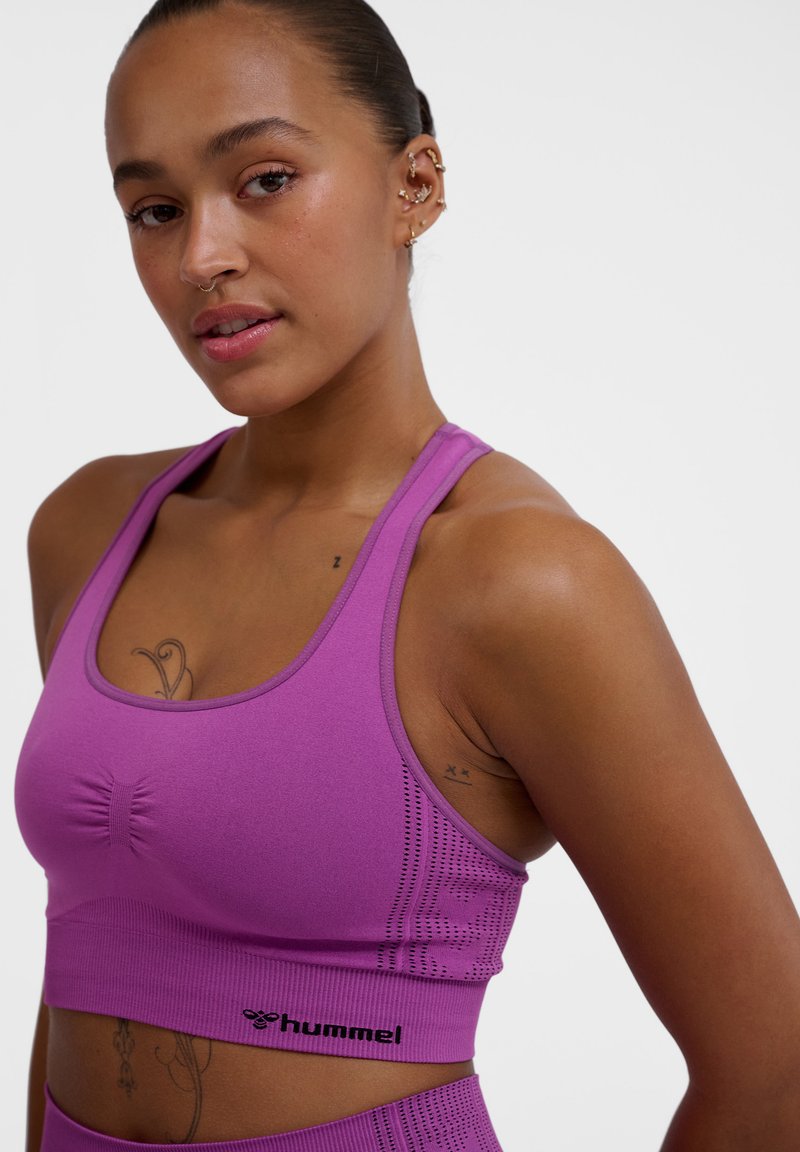 Fioletowy sportowy top w wersji crop z ribbowanej tkaniny, z wyciągniętym przodem i logo na dolnej krawędzi, zaprojektowany z myślą o komforcie i wsparciu.