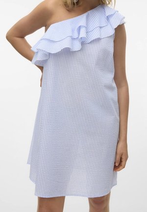 Femme portant une robe rayée bleu clair et blanc à une épaule avec un détail à volants sur la poitrine, debout devant un fond blanc.