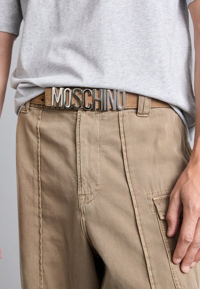 Pruun nahast vöö, millel on metallik hõbedane "MOSCHINO" logo pannal, kantud beežide pükstega ja halli T-särgiga.