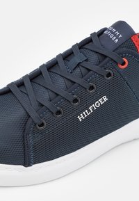 Marineblaue Sneaker aus Mesh-Material, weiße Gummisohle, auffälliges Schnürsystem und rote Akzente. Mit "HILFIGER" Branding an der Seite.