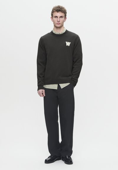 Suéter de punto oscuro con cuello redondo que presenta un logotipo de letra "W" blanca, superpuesto a una camiseta de rayas verdes y blancas, combinado con pantalones negros.