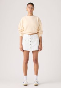 Νέα γυναίκα που φοράει μπεζ φούτερ, μίνι φούστα με κουμπιά μπροστά και τσέπες σε λευκό χρώμα, λευκές κάλτσες και λευκά αθλητικά παπούτσια με μπλε και κόκκινες ρίγες.