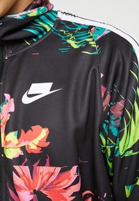 Svart jacka med livfulla blommönster i rosa, grönt och blått. Har en dragkedjestängning och Nike-logotyp på bröstet och axelsömmen.