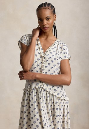 Moteris su pintais plaukais, vilkinti baltą su mėlynais gėlių raštais ir raukiniais puoštą suknelę, pozuojanti su ranka prie kaklo neutraliame fone.