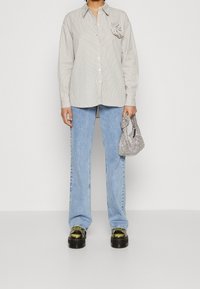 Gestreiftes Hemd mit floralen Akzenten, helle Denim-Jeans im Schlag, und eine gemusterte Mini-Handtasche. Klobige schwarze Schuhe mit metallischem Finish.