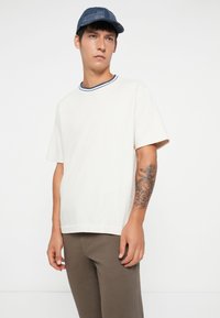 White short-sleeve t-shirt with a blue-striped neckline, paired with brown pants. The person is wearing a dark blue cap and shows a tattoo on the arm.