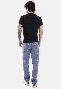 Homme aux cheveux bouclés et tatouages sur les bras, portant un t-shirt noir, un jean bleu et des baskets blanches, debout de dos.
