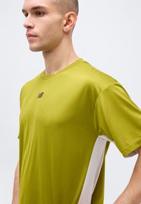 Άνδρας με κοντά μαλλιά που φοράει αθλητική μπλούζα New Balance σε πράσινο λάιμ με λευκές πλέξεις στα πλαϊνά, κοιτάζει προς τα αριστερά.