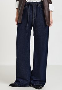 Jeans a gamba larga in denim blu scuro, con una vita con coulisse, cuciture a contrasto e una superficie morbida. Abbinati a una giacca di pelle marrone.