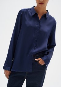 Granatowa satynowa koszula z długim rękawem, zapinana na guziki z klasycznym kołnierzykiem i lekko luźnym krojem, zestawiona z ciemnymi dżinsami.