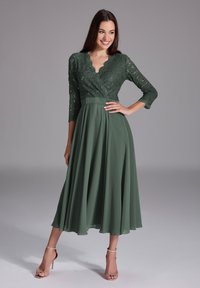 Robe verte avec des manches trois-quarts en dentelle, décolleté festonné, corsage ajusté et jupe fluide. Associée à des sandales à talons hauts avec des brides.