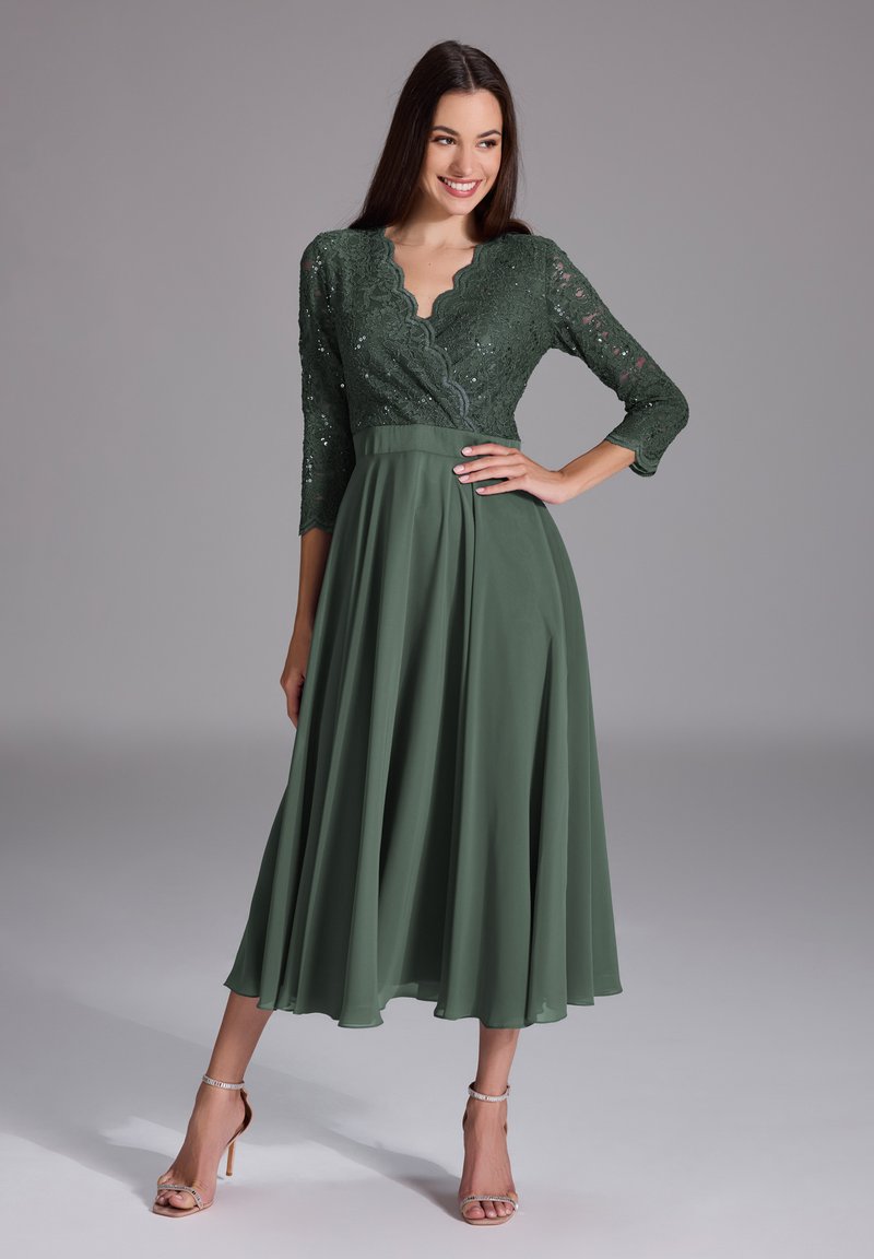 Robe verte avec des manches trois-quarts en dentelle, décolleté festonné, corsage ajusté et jupe fluide. Associée à des sandales à talons hauts avec des brides.