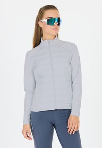 Lys grå zip-jakke med pufforfrontpaneler, lange ærmer og høj krave. Båret med blå træningsleggings og reflekterende solbriller.