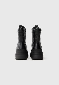 Botas de tornozelo em couro preto com sola de borracha preta robusta, zíperes traseiros e acentos de logótipos proeminentes no calcanhar e na alça.