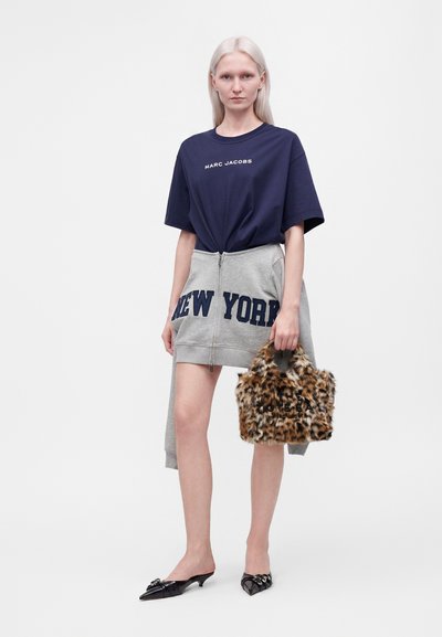 Femme portant un t-shirt Marc Jacobs bleu marine, une jupe grise zippée avec l'inscription New York, tenant un sac à main imprimé léopard et des chaussures noires pointues.