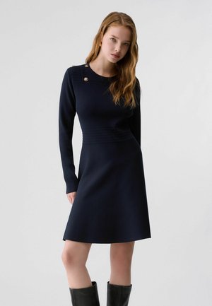 Femme aux longs cheveux ondulés portant une robe bleu marine à manches longues avec des boutons dorés aux épaules et à la taille, associée à des bottes noires montantes jusqu'aux genoux.
