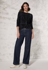Schwarzer gestrickter Pullover mit geometrischem weißen Muster, kombiniert mit weit geschnittenen dunkelblauen Jeans und weißen Turnschuhen. Glatte Textur, lässige Passform.