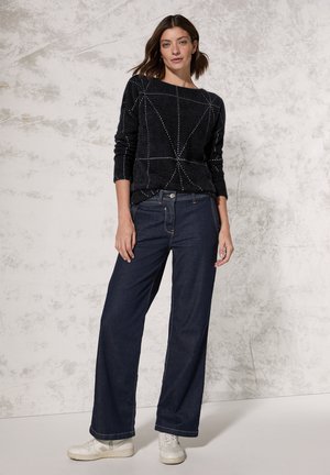 Sort strikket sweater med geometrisk hvidt mønster, parret med brede mørkeblå jeans og hvide sneakers. Glat tekstur, afslappet pasform.