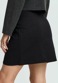 Mini jupe noire en tissu extensible, dotée d'une silhouette ajustée et de coutures nettes. Associée à un blazer structuré gris.