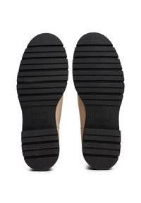 Une paire de chaussures beiges avec des semelles en caoutchouc noir striées affichant le logo "Tommy Jeans" au niveau du talon, vue de dessous.