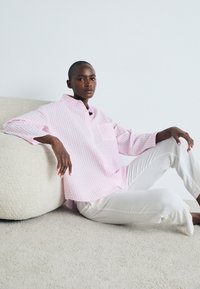 Ροζ και λευκό καρό πουκάμισο με τσέπη στο στήθος, συνδυασμένο με λευκά παντελόνια, φορεμένο από ένα μοντέλο που κάθεται σε ένα υφή φωτεινού φόντου.