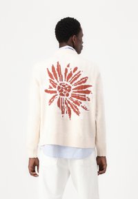 Gilet crème avec un grand motif floral rouge dans le dos. Texture tricotée, poignets côtelés et coupe décontractée sur une chemise à col.