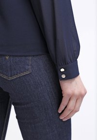 Manche de blouse bleu marine avec deux boutons argentés sur le poignet, associée à un jean denim bleu foncé, montrant la poche arrière et une main reposant sur le côté.