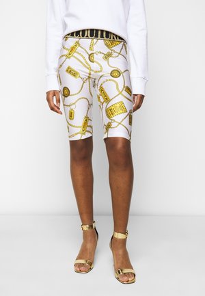 Witte biker shorts met hoge taille, gouden ketting- en tagprint, zwarte tailleband met gouden tekst, gedragen met een wit top met lange mouwen en gouden hakken.
