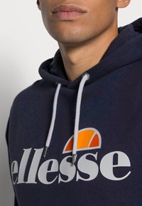 Marinblå hoodie med vita dragsnören och orange logotypdetalj. Har mjukt tyg, avslappnad passform och ett stort tryckt varumärkesnamn.