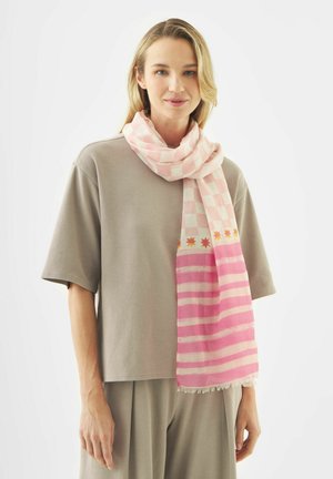 Junge Frau trägt ein beiges, locker sitzendes T-Shirt und eine Hose sowie einen rosa-weiß karierten Schal mit Stern- und Streifenmustern.