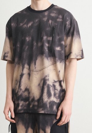Unisex schwarz-beiges Batik-T-Shirt mit vertikaler Reißverschlusstasche auf der Brust, getragen mit passenden Batik-Hosen.