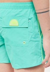 Shorts de bain turquoise en matériau léger, dotés d'une poche arrière avec un logo jaune, des coutures d'accent et un petit détail de grommet.